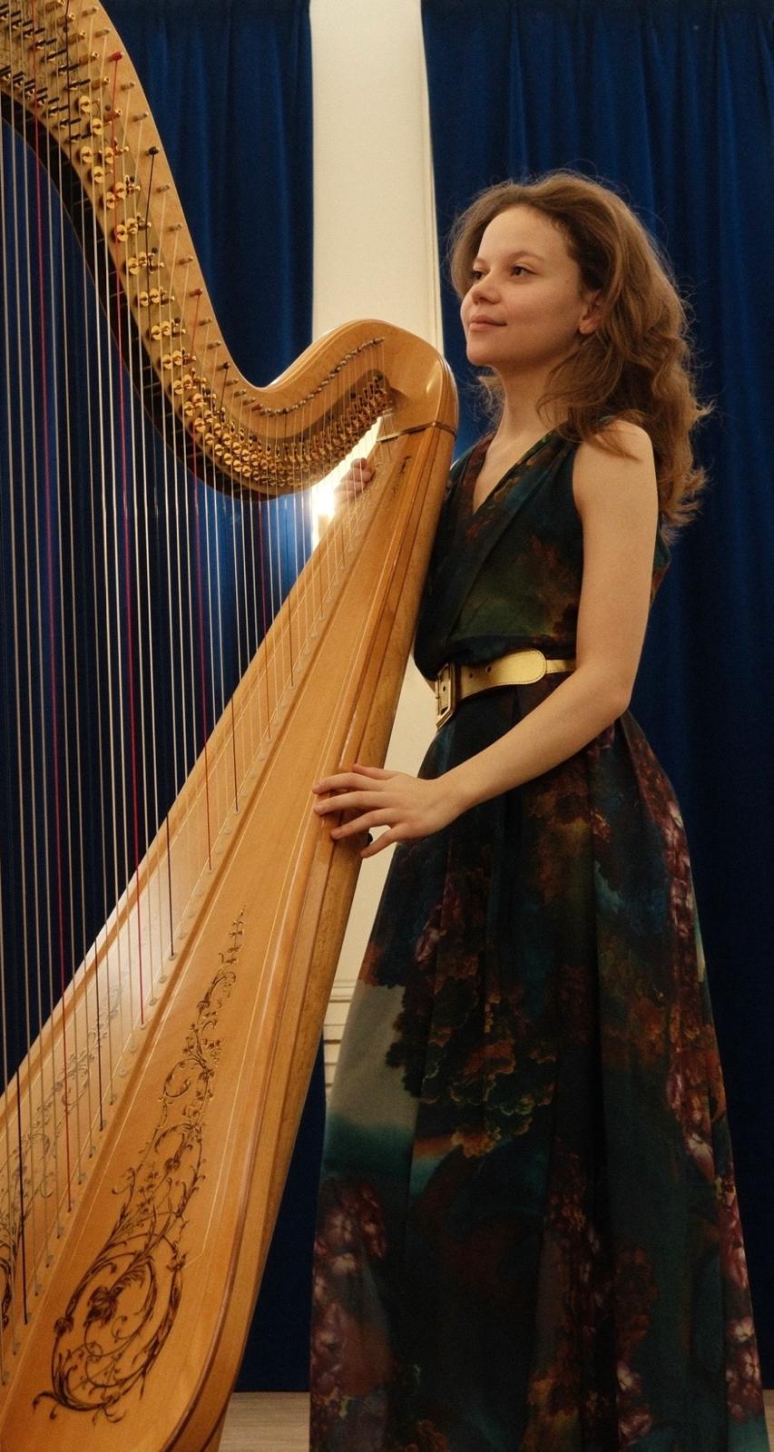 Une jeune musicienne se tenant debout près d'une harpe en bois, vêtue d'une robe élégante aux motifs colorés.