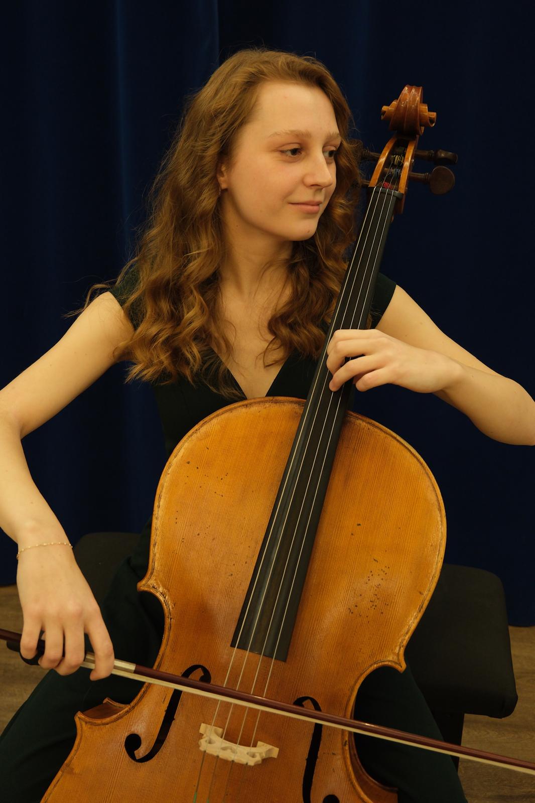 Une jeune musicienne joue du violoncelle, les cheveux bouclés et un sourire concentré, avec un fond bleu.